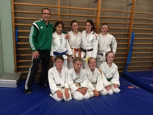 Gruppenspiel von einem Trainer und sieben jungen Judoka in weißen Gi auf einer Judomatte vor einer Wand mit Klettergerüst.