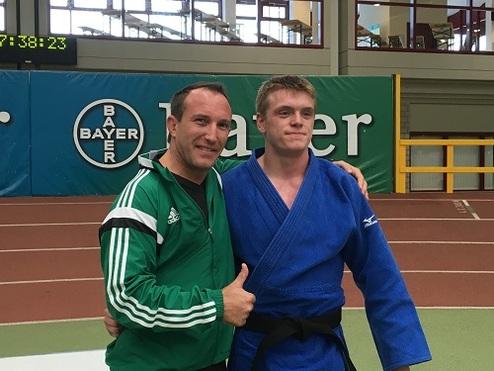 Zwei Männer posieren zusammen auf einer Judomatte, einer trägt einen blauen Judogi, der andere ein grünes Trainingsoutfit.