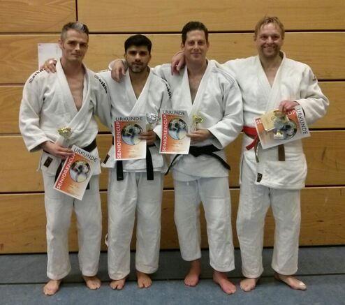 Vier Judokas in weißen und farbigen Judogi stehen zusammen und halten Pokale und Urkunden nach einem Wettkampf in der Halle.