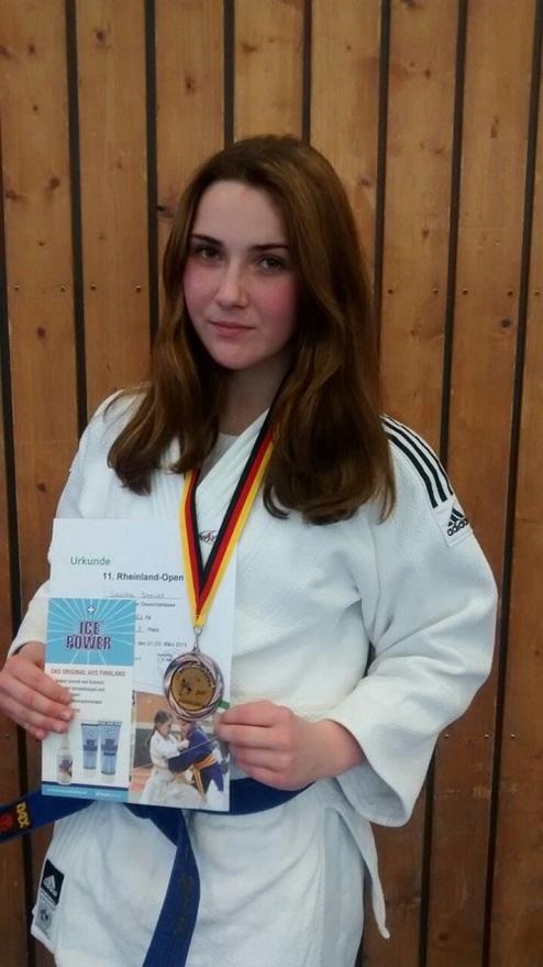 Junge Judoka in weißem Gi hält Medaille und Urkunde des Rheinland-Open, Hintergrund aus Holzlatten.