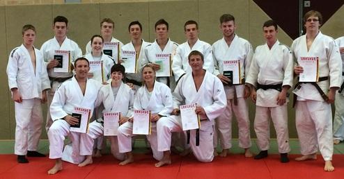 Gruppe von Judokas in weißen Anzügen, einige halten Urkunden, in einer Sporthalle auf einer Matte.