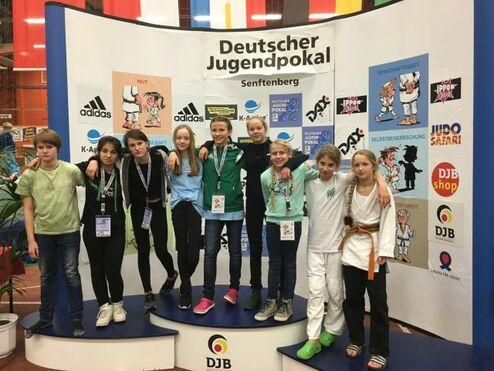 Gruppe von Kindern und Jugendlichen auf dem Siegertreppchen beim Deutschen Jugendpokal in Senftenberg, umgeben von Werbebannern.