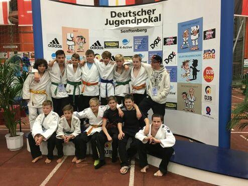 Gruppenscreenshot von Judokas in weißen und farbigen Anzügen vor einem Banner des Deutschen Jugendpokals in Senftenberg.