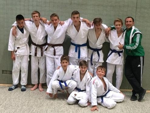 Gruppe von Jungen in weißen und blauen Judoanzügen lächelt vor einem grauen Hintergrund, umarmt von einem Trainer.
