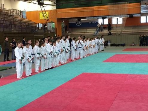 Gruppe von Judoka in weißen Anzügen steht auf einer Turnmatte in einer Sporthalle, bereit für den Wettkampf.