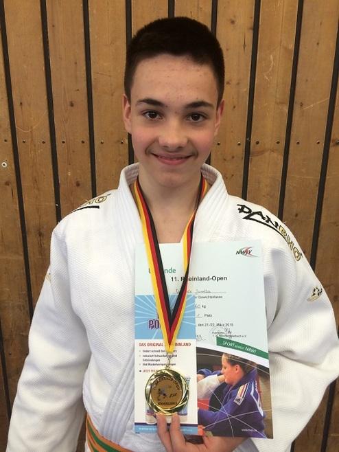 Junge in einem Judoanzug mit Medaille und Urkunde vom Rheinland-Open, vor einer Holzwand.