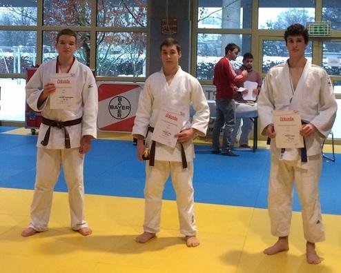 Drei Judoka in Weiß und Braun halten Urkunden in einer Sporthalle mit blauen und gelben Matten.
