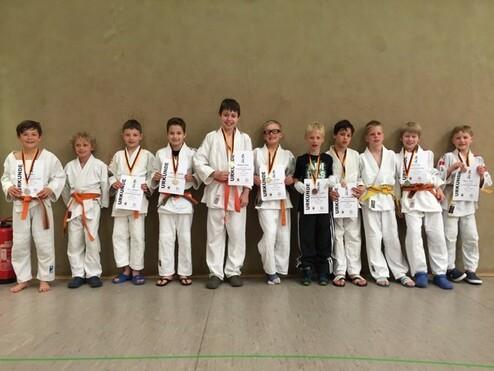 Gruppe von zehn Kindern in Judogi mit Medaillen, die stolz ihre Urkunden in einer Sporthalle präsentieren.