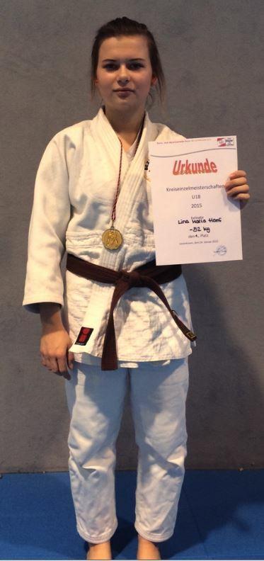 Junge Judoka in weißem Gi mit braunem Gürtel hält Urkunde und Medaille nach erfolgreicher Teilnahme an Meisterschaft.