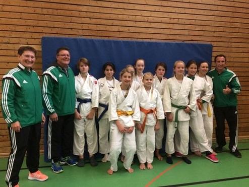 Gruppenbild mit Judokas in weißen und farbigen Gürteln sowie Trainern in grünen Sportanzügen, vor einer Holzwand.