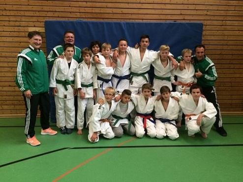 Gruppennfoto von Judokas in weißen und bunten Anzügen, umgeben von Trainern in grüner Sportbekleidung in einer Halle.