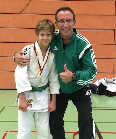 Junge in Judogewand mit Medaille um den Hals, posiert mit Trainer in Sportkleidung in einer Turnhalle.