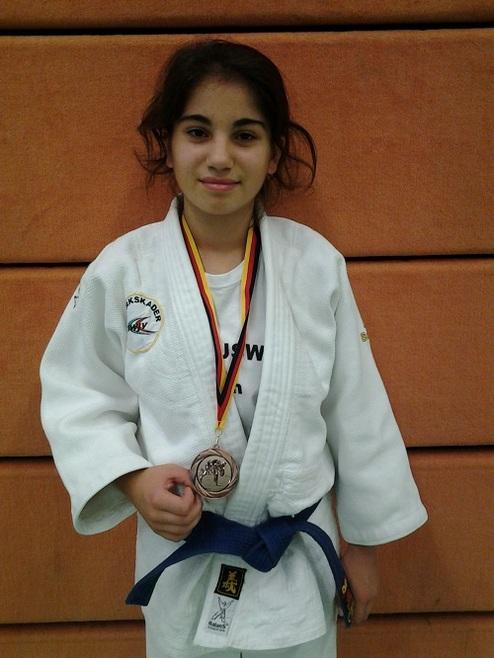 Mädchen in Judogi mit blauer Gürtelfarbe hält eine Medaille in der Hand, Hintergrund in Orange.