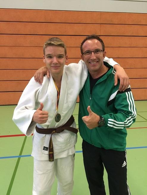 Zwei Männer in Judokleidung, lächeln und zeigen Daumen hoch, in einer Sporthalle mit grüner Matte.