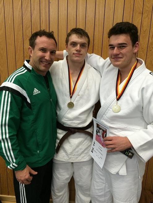 Drei Männer in Judogewändern mit Medaillen posieren lächelnd vor einer Holzwand. Ein Mann hält ein Zertifikat in der Hand.