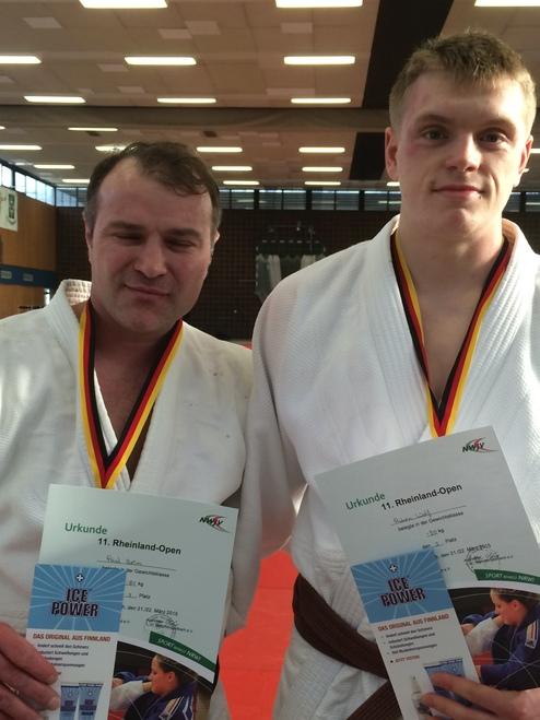 Zwei Judoka mit Medaillen in weißen Judogis halten Urkunden des Rheinland-Open-Turniers in einer Sporthalle.