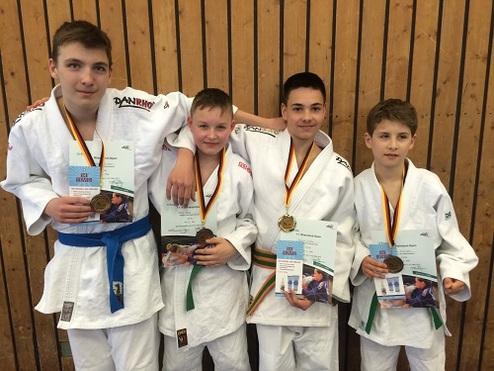 Vier jugendliche Judoka in Uniform stehen vor einer Holzwand, halten Medaillen und Urkunden in der Hand.