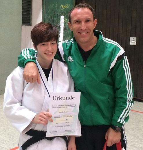 Junge mit Kurzhaarschnitt in Judogi hält eine Urkunde, umarmt von einem Trainer in Sportkleidung.