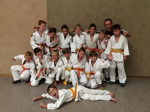 Gruppenzusammenkunft von Kindern in Judoanzügen mit gelben und orangen Gürteln, lächelnd vor einer Wand.