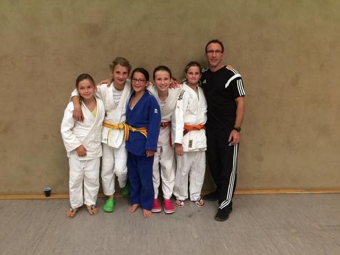 Sechs Kinder in Judogis und ein Trainer posieren lächelnd vor einer Wand in einer Sporthalle.