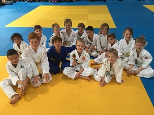 Gruppe von 15 Kindern in Judogis, sitzend auf einer gelben und blauen Judomatte, lächelnd und in zwei Reihen angeordnet.