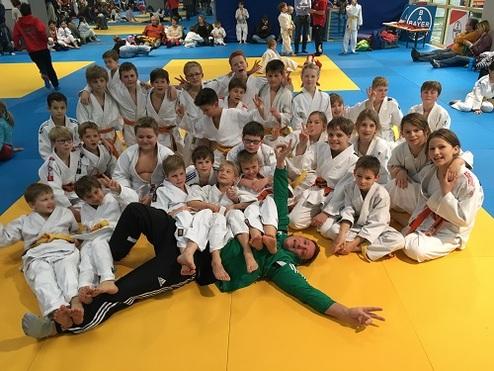 Gruppenzusammenkunft von Kindern in weißen Judogis auf einer blauen Judomattenfläche, teils mit gelben Gürteln.