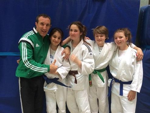 Gruppenselfie von vier Judoka in Weiß und Grün mit Trainer in Sportjacke vor einer blauen Mattenwand.