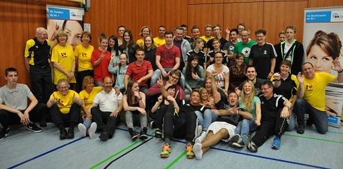 Gruppenfoto einer Tischtennisveranstaltung mit Teilnehmern in verschiedenen T-Shirts, lächelnd auf einer Sporthalle.