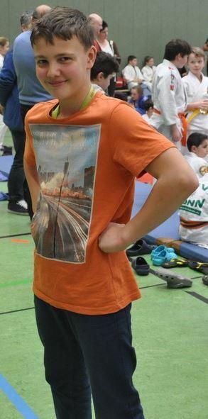 Jungen in orangefarbenem T-Shirt mit Stadtmotiv steht selbstbewusst in einer Sporthalle mit anderen Kindern im Hintergrund.
