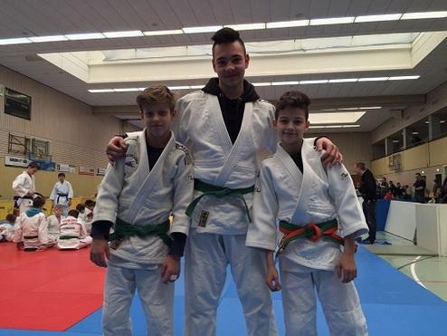 Drei Jungen in Judoanzügen posieren freundlich auf einer Judomatte in einer Sporthalle.