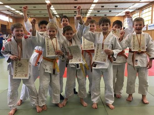 Gruppe von jugendlichen Judoka in weißen Judogi mit Medaillen und Urkunden, glücklich posierend nach einem Wettkampf.