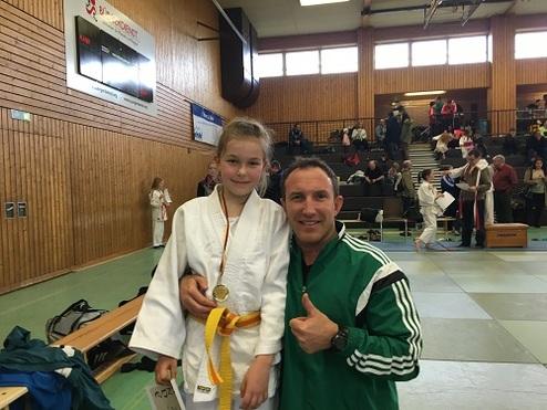 Junge mit Medaille und Gürtel umarmt Trainer in Sporthalle während eines Judoturniers. Zuschauer im Hintergrund.