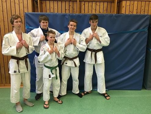 Fünf Judokas in weißem und braunem Gi stehen freundlich vor einer blauen Wand in einer Sporthalle.