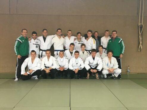 Gruppe von 16 Judokas in weißen und schwarzen Judogis, versammelt auf einer Judo-Matte in einem Sportraum.