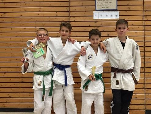 Vier Kinder in Judogi stehen freundlich zusammen, halten Pokale und Urkunden in der Hand, vor einer Holzwand.