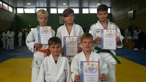 Fünf Kinder in Judo-Anzügen halten Urkunden in einer Sporthalle mit Judo-Matten im Hintergrund.