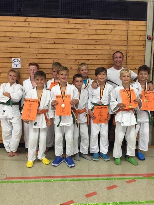 Gruppe von zehn Jungen in Judo-Anzügen, stolz mit Medaillen und Urkunden vor einer Holzwand.