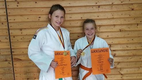 Zwei Mädchen in Judokas zeigen stolz ihre Urkunden und Medaillen vor einer Holzwand.