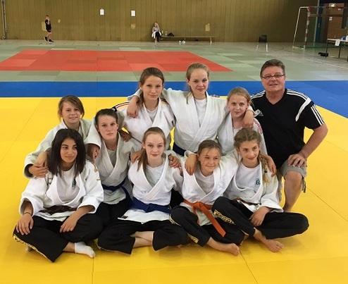 Gruppenfoto von neun Judoka in Gis, umgeben von einer gelben Matte; ein Trainer sitzt mit ihnen auf dem Boden.