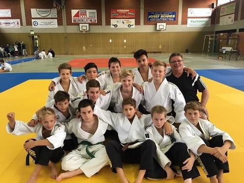 Gruppe von Jugendlichen in Judoanzügen posiert auf einer Judo-Matte, umgeben von weiteren Judo-Praktizierenden.