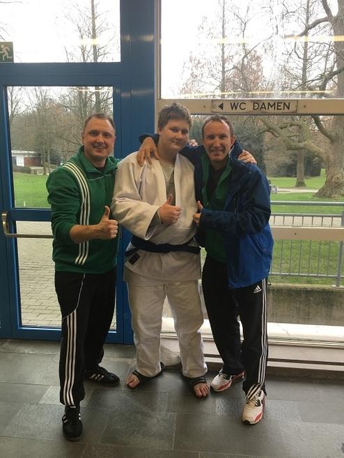 Drei Personen stehen vor einer Fenstertür und posieren im Judogi, zwei mit Daumen nach oben, lächeln.