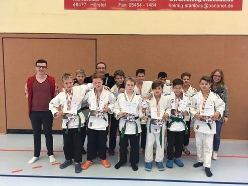 Gruppe von Jungen in Judoanzügen mit Medaillen und Urkunden, umringt von drei Erwachsenen in einer Sporthalle.