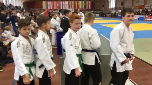 Fünf Kinder im Judoanzug stehen wartend in einer Schlange bei einem Turnier, im Hintergrund ist eine Sporthalle zu sehen.