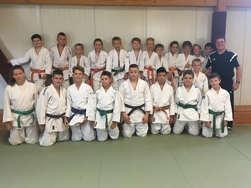 Gruppierung von 20 Judokas in weißen, grünen und orangenen Judogis in einer Sporthalle, begleitet von einem Trainer.