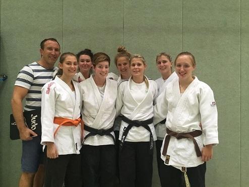 Gruppe von sieben Judokas in weißen und schwarzen Judogis, posierend mit einem Trainer in einer Turnhalle.