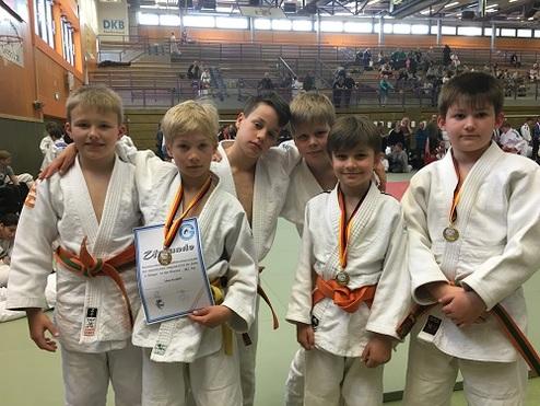 Fünf Kinder in Judoanzügen posieren stolz mit Medaillen und einem Zertifikat in einer Sporthalle.