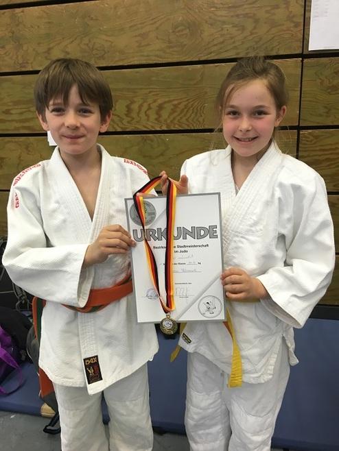 Zwei Kinder in Judokleidung halten stolz eine Urkunde mit Medaille nach einem Wettkampf.