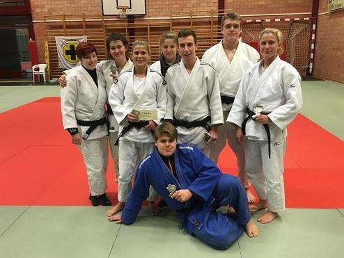 Gruppe von Judoka in Sportanzügen posiert auf der Matte nach einem Wettkampf, mit Medaillen und Pokal in der Hand.