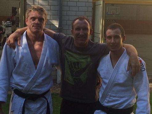 Drei Männer in Judoanzügen stehen lächelnd nebeneinander in einem Freiluftbereich.