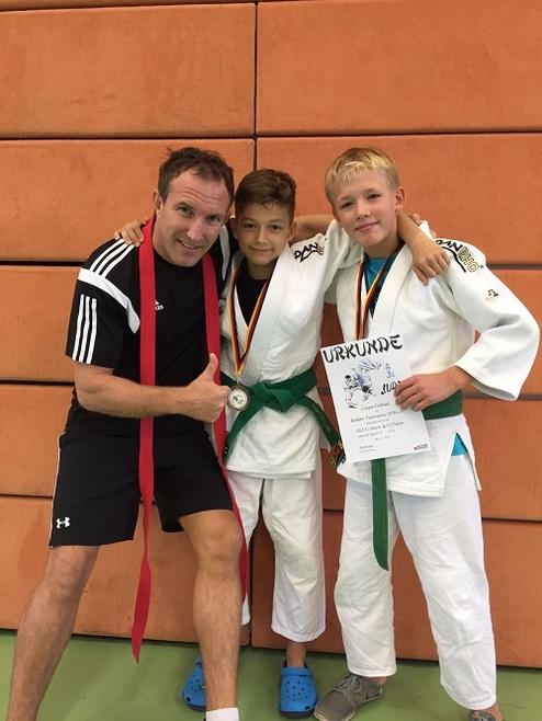 Trainer posiert mit zwei Judoka in weißen Anzügen, die Medaillen tragen und eine Urkunde halten.